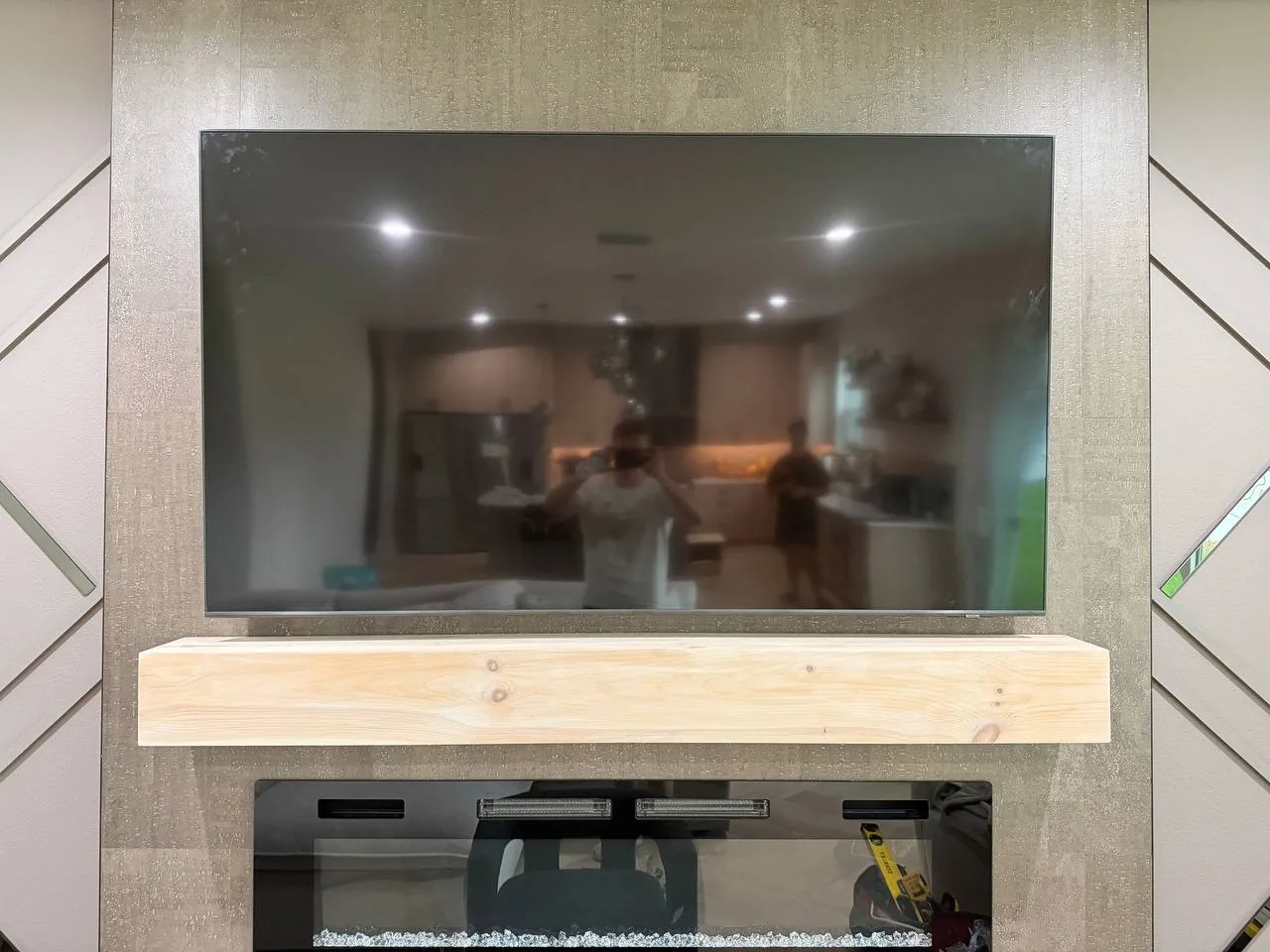 A wide TV mounted on a textured feature wall above a floating light-wood mantel shelf and an electric fireplace.