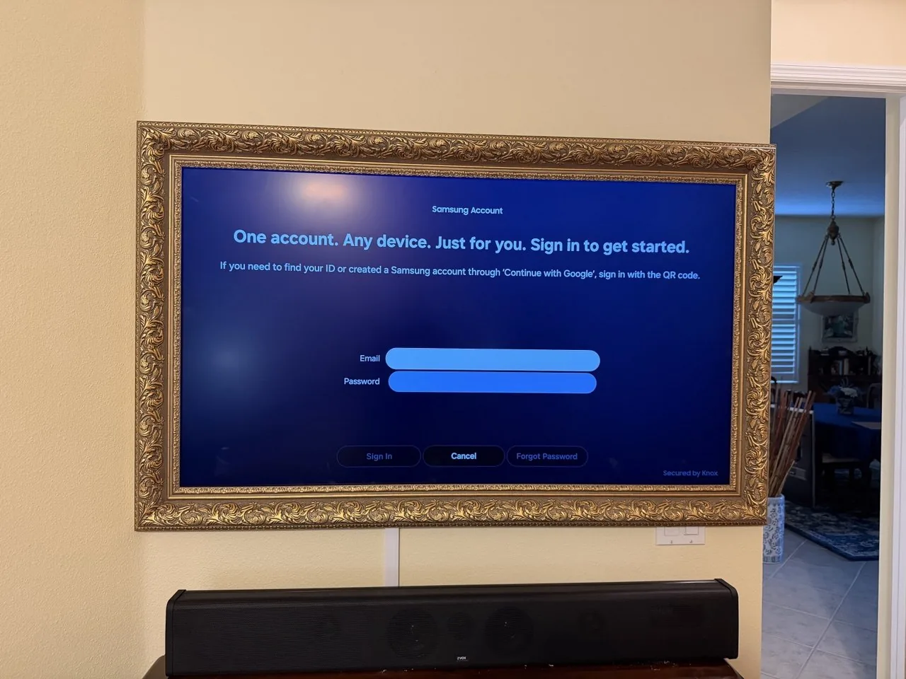 A TV in a decorative gold picture frame mounted on the wall above a soundbar, with the cable concealed in a white raceway.